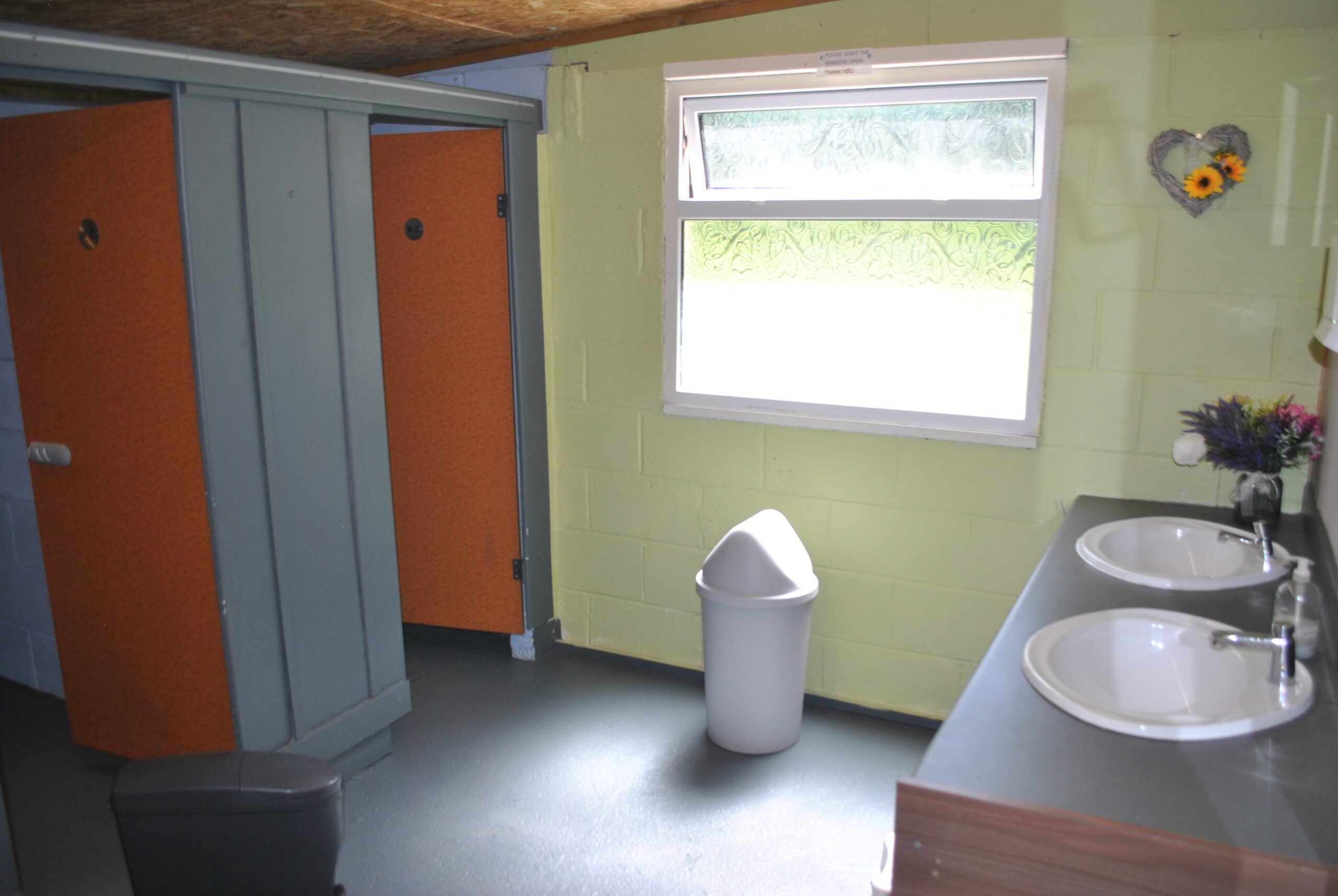 2 wash basins, toilet door and waste bin with a large frosted window in the background